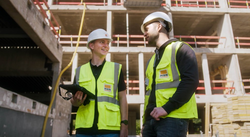 Zwei Personen schauen den digitalen Bauablaufplan an. Im Hintergrund sieht man die Baustelle im Hochbau.