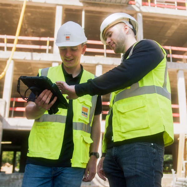 Bauleiter und specter Mitarbeiter sprechen auf der Baustelle über die Bausoftware Vorteile. Dabei bedient der specter Mitarbeiter den 3D-Plan auf den Tablet. Im Hintergrund ist die Baustelle im Hochbau zu sehen.