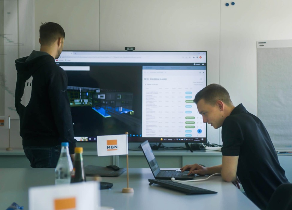 Im Besprechungsraum des Generalunternehmen sind zwei Personen mit der digitalen Baudokumentation durch specter beschäftigt. An der Wand ist ein großer TV zur Präsentation für digitale Baudokumentation.