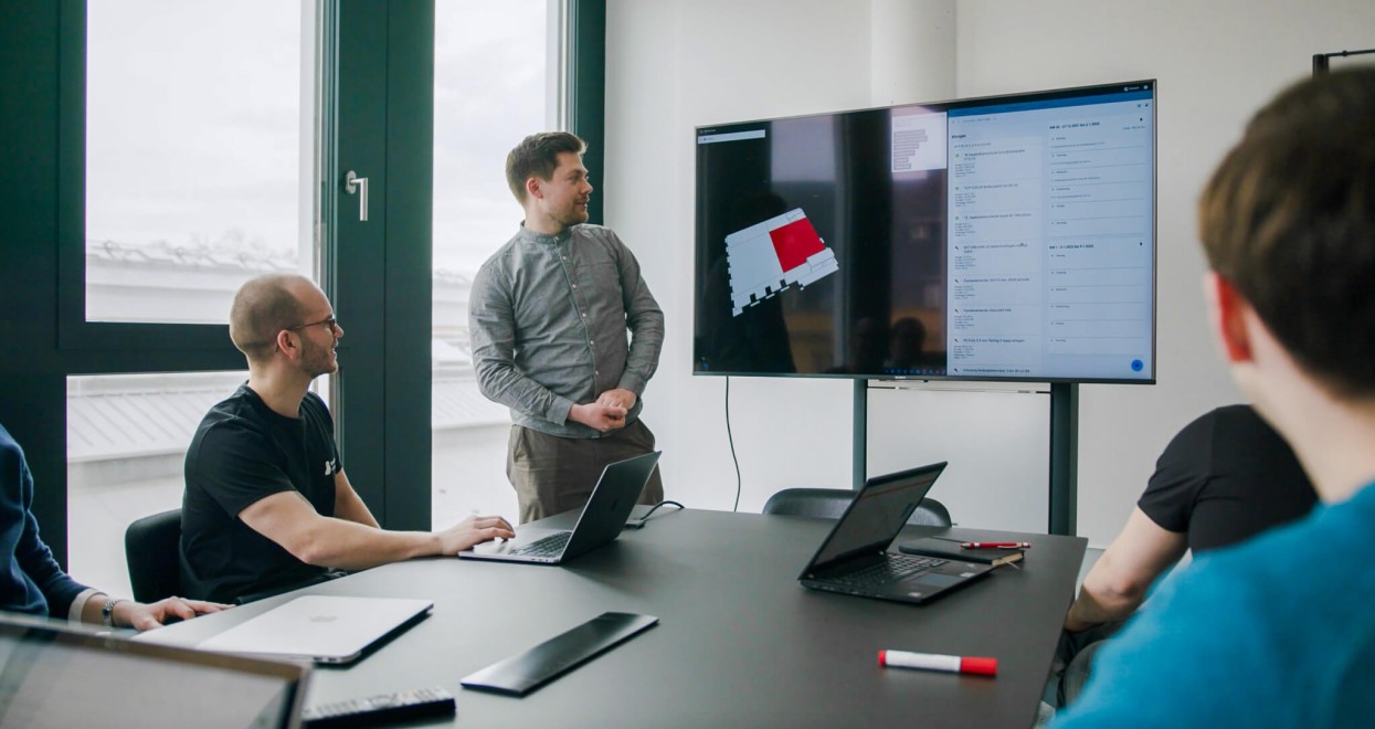 Der digitale Baufortschritt für Projektbeteiligte wird im Besprechungsraum besprochen. Am Bildschirm wird die Bausoftware für Projektbeteiligte besprochen und mehrere Personen nehmen Teil.