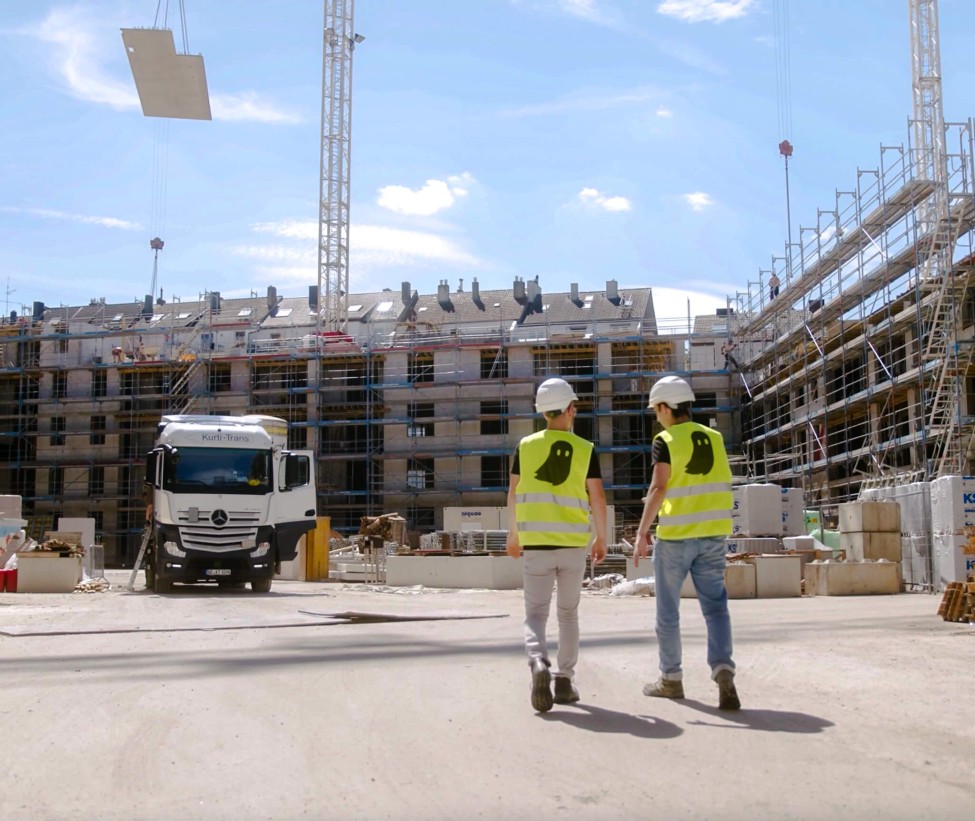 Die Hochbau Baustelle ist in der Sonne zu sehen. Zwei Personen in specter automation Warnweste laufen über den Platz und sprechen über digitalen Baufortschritt.