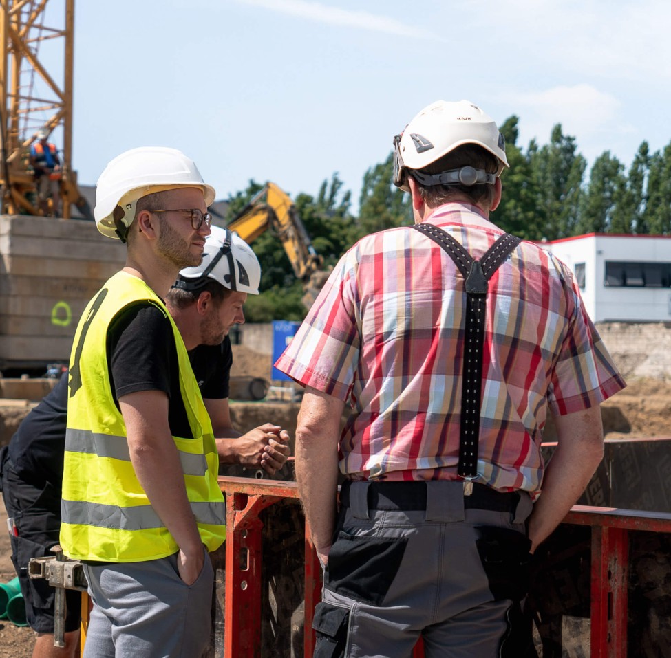 News der Baubranche werden direkt auf der Baustelle besprochen. Drei Personen sind auf der Baustelle in der Sonne und tragen Sicherheitskleidung.