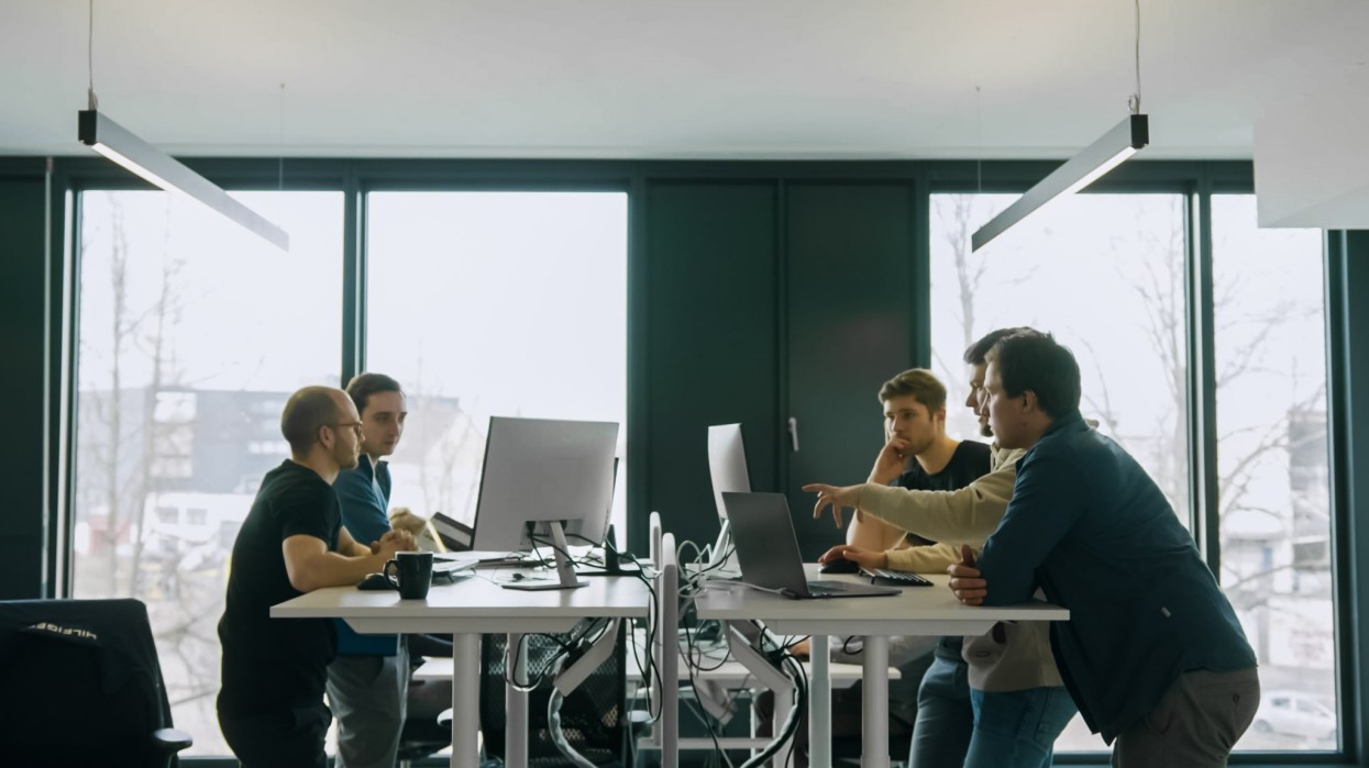 News der Baubranche werden direkt von einer Personengruppe am Arbeitsplatz aufgerufen. Fünf Personen stehen im offenen Bereich und schauen auf mehrere PC.